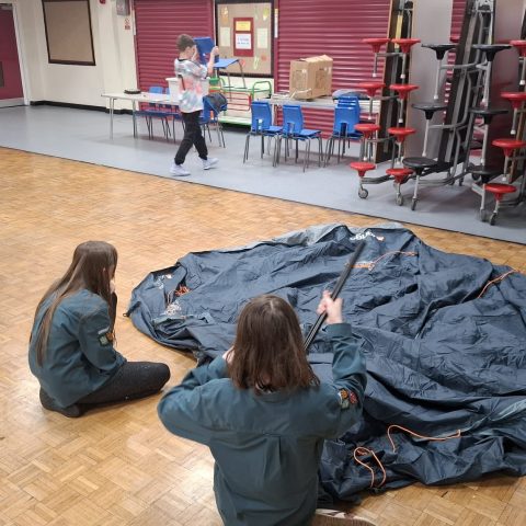 Scouts laying out a new tent on the hall floor and fitting the poles